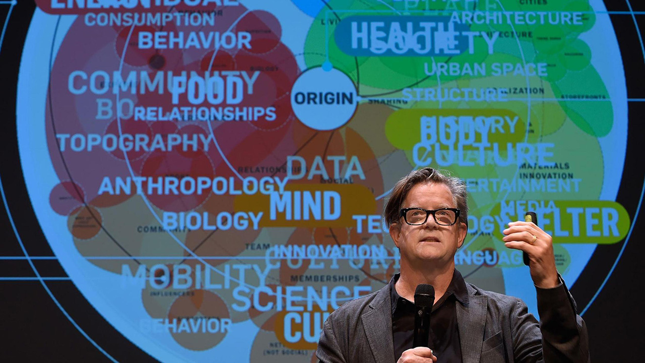 Alex McDowell talking on stage, holding a microphone, with projected behind him a graphic showing a world building brainstorm technique