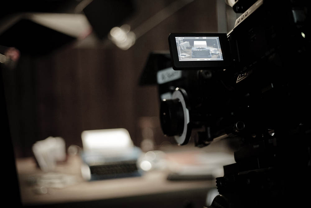 Behind the scenes on The Black Diamond shoot: a cinema camera on a rig is focused on a dimly lit desk with a vintage typewriter in the background, captured in a dark studio.