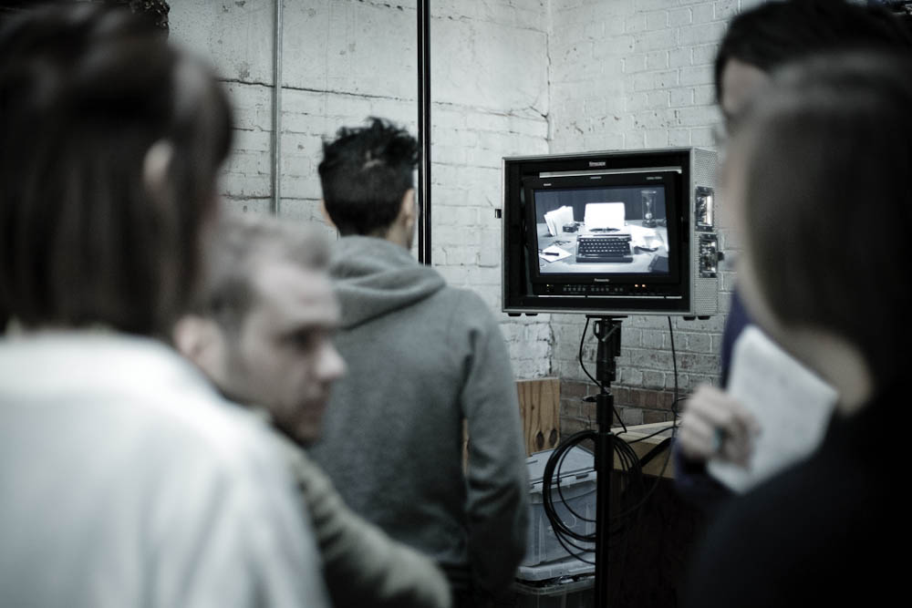 Behind the scenes on The Black Diamond shoot: crew members gather around a Panasonic broadcast monitor displaying a black-and-white typewriter scene, against a white-painted brick wall.