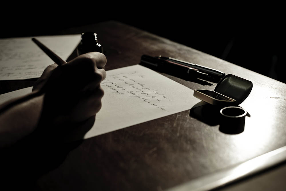 Behind the scenes on The Black Diamond shoot: a hand writes by lamplight on paper, with fountain pens and an ink bottle arranged on a steel surface, styled in the noir atmosphere of the film.