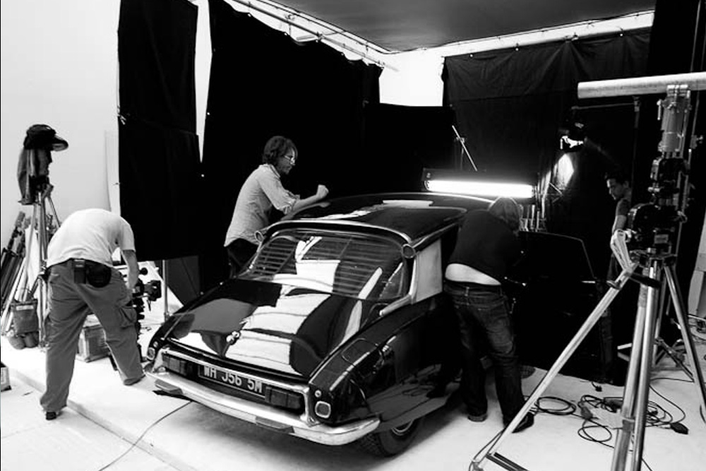 Behind the scenes on The Black Diamond shoot: a black vintage car with a British number plate is surrounded by crew members in a large white studio space, with multiple lights and camera rigs visible.