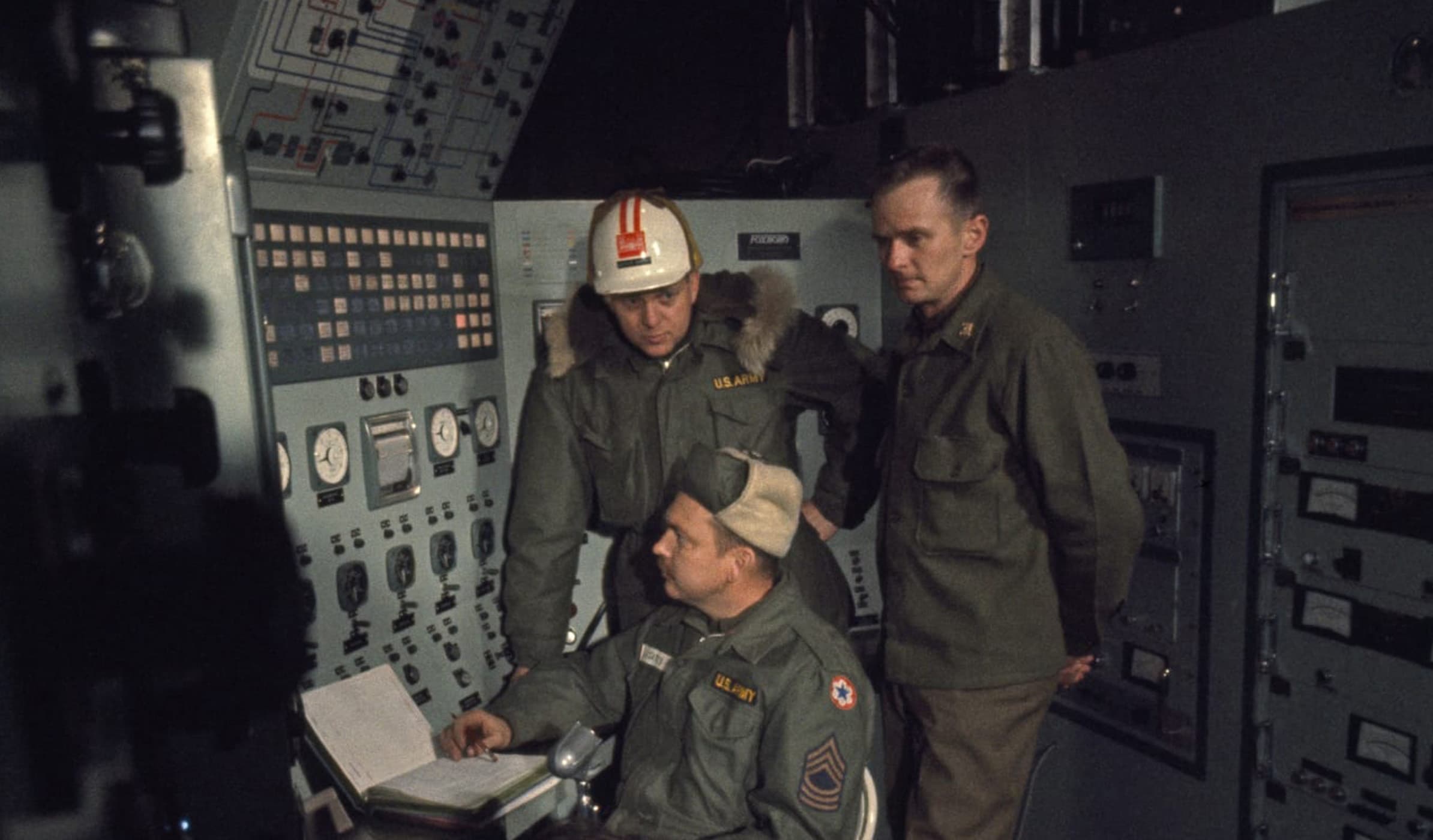 Three US Army personnel gathered in front of a control panel within Camp Century. One individual is attentively focused on the panel, while the others are observing his actions.