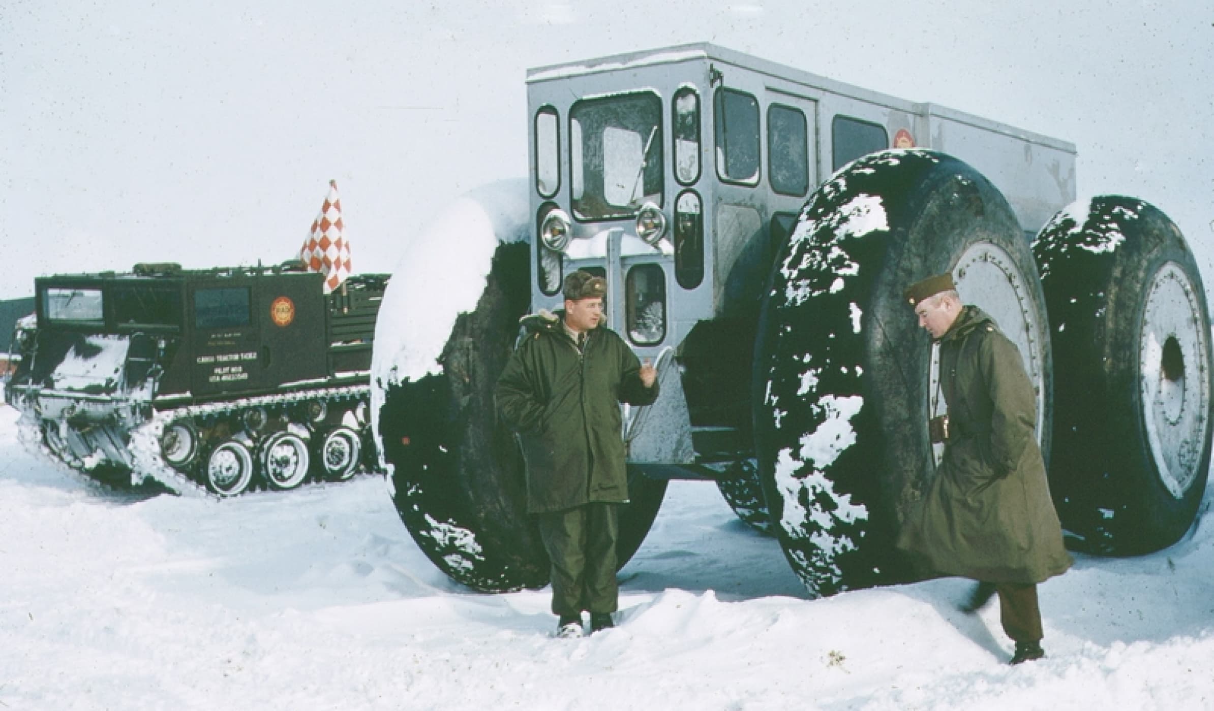 Two US Army personnel stand in the Arctic snow in front of a snow-adapted vehicle, notable for its oversized tires towering above the men.