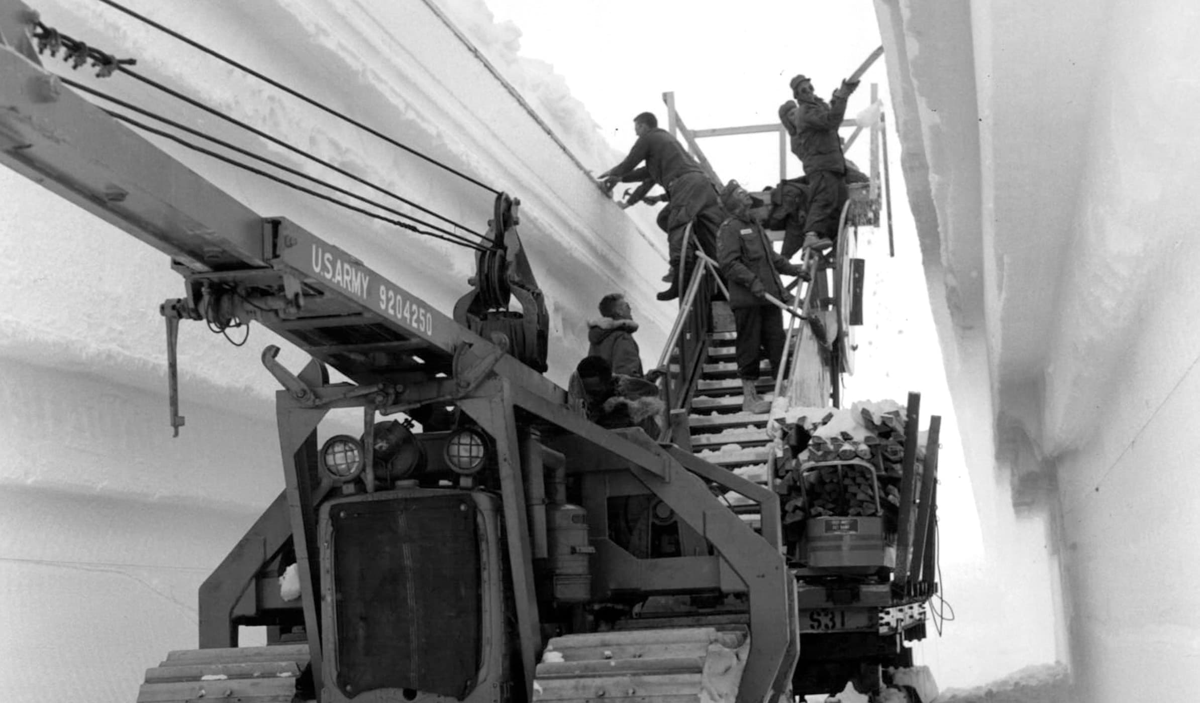 Seven men on a US Army aerial work platform: one operates the crane, four work on carving a tunnel through deep snow, and two oversee the operations.