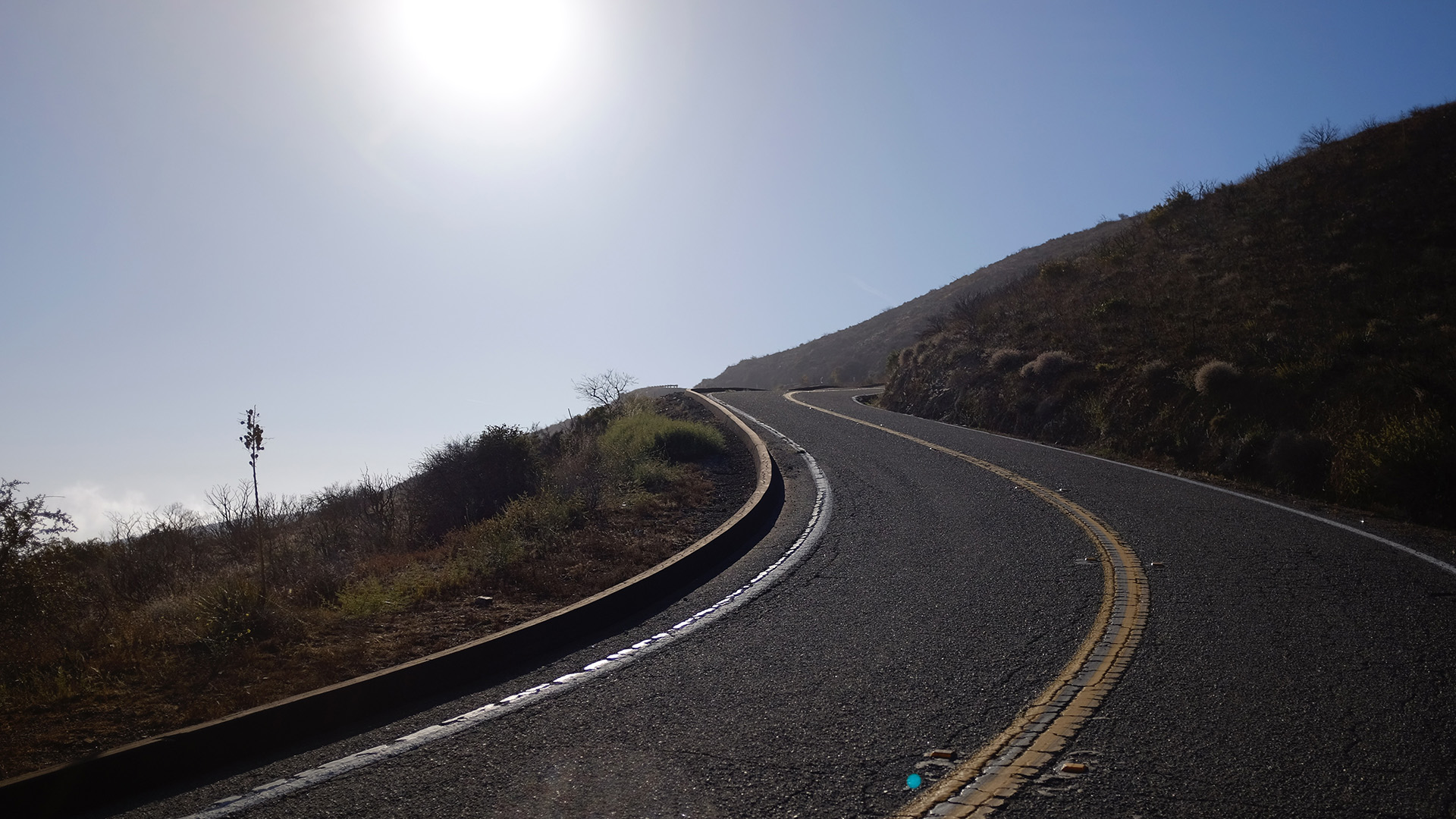 A deserted winding road climbs a dry Malibu hillside under a blazing sun, shot from road level looking uphill.