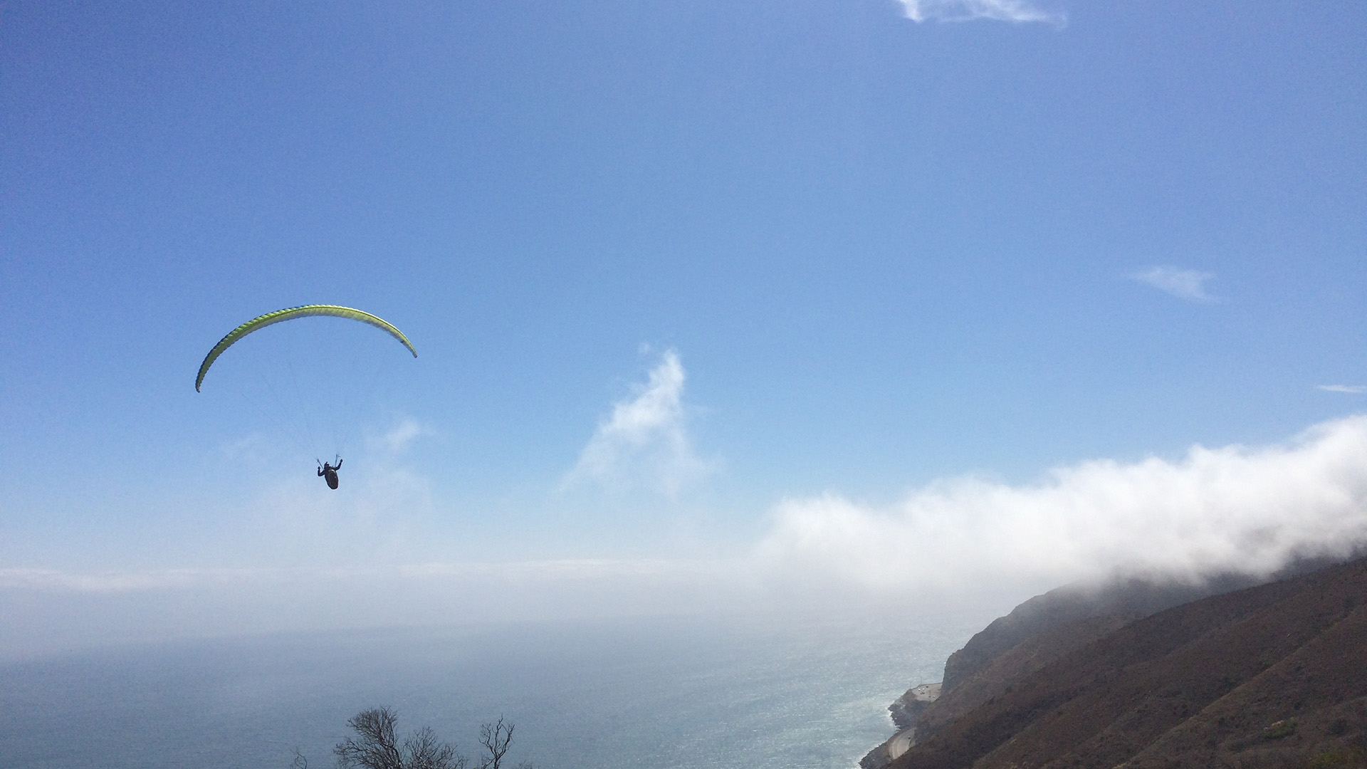 A paraglider soars above the Malibu coastline, with low clouds hugging the ocean and coastal cliffs below.