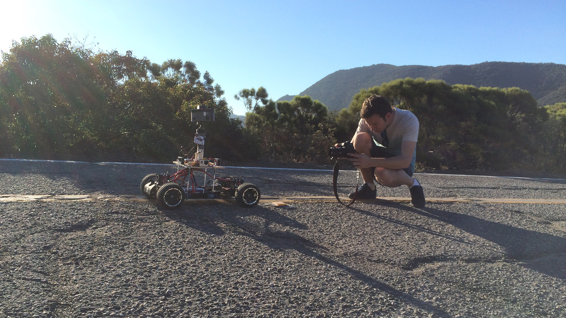 Behind the scenes on the Elevate shoot: a crew member crouches to adjust a custom-built RC car carrying a camera rig on a Malibu road, mountains visible behind.