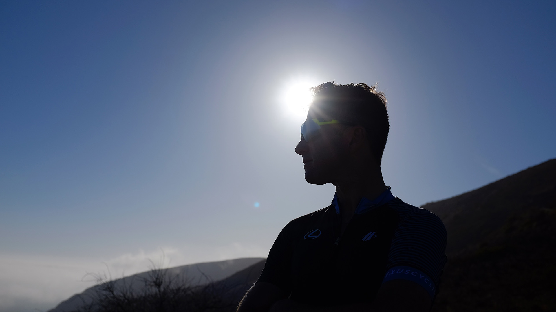 Silhouette of cyclist Christian Vande Velde in profile against a bright Malibu sky, the sun haloed behind his head, wearing a Lexus cycling kit and sunglasses.