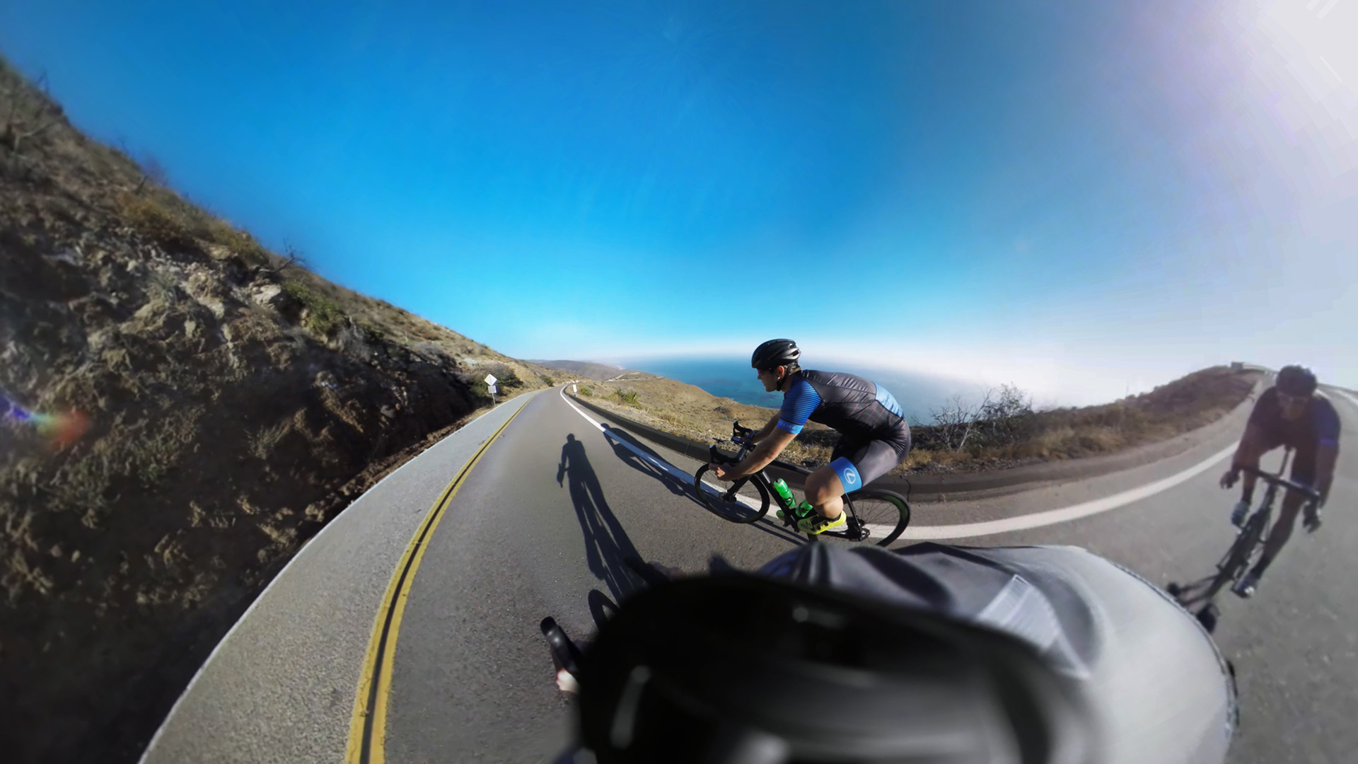 Elevate: Christian Vande Velde in his Lexus cycling kit leans into a curve on a Malibu coastal highway, the Pacific Ocean and coastal cliffs stretching to the horizon behind him, with a camera rig mount visible in the foreground.