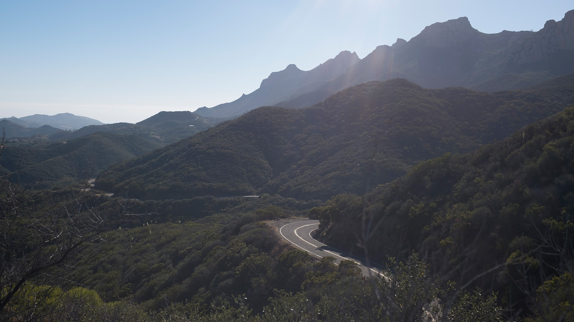 Elevate: a winding two-lane road cuts through the forested green hills of Malibu, with jagged rocky peaks catching sun rays in the background.