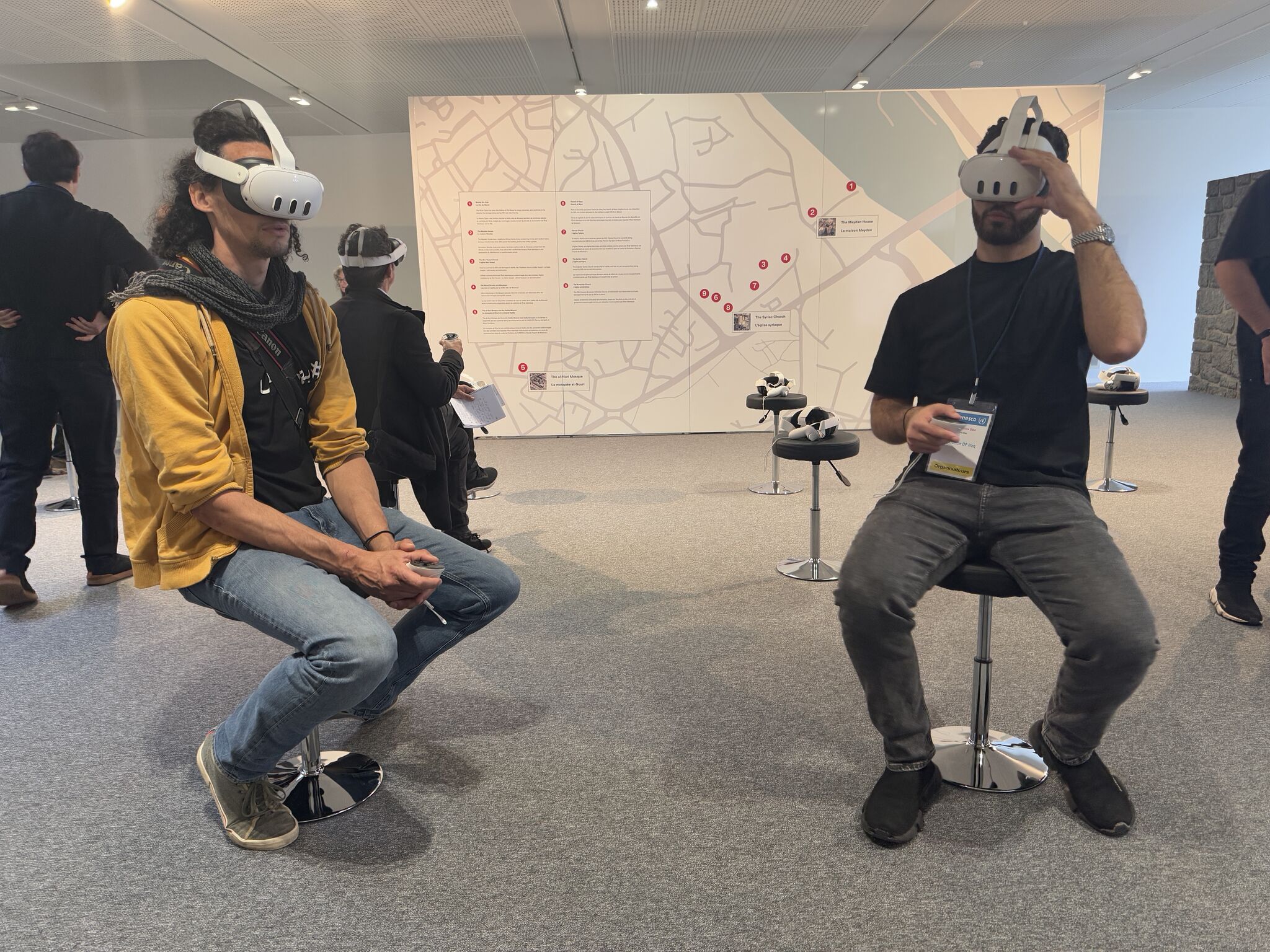 Two people sit on stools wearing VR headsets in front of a map of Mosul