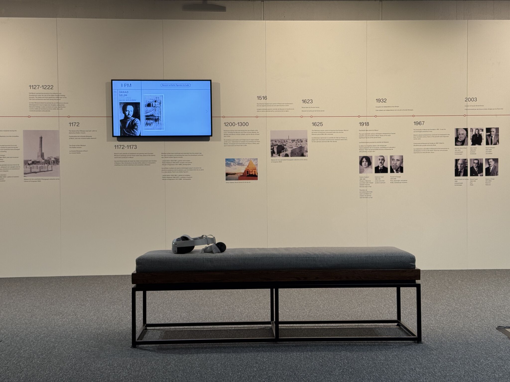 A wall featuring a timeline of old Mosul with a screen, images and text. In front of it is a bench with a VR headset on it.