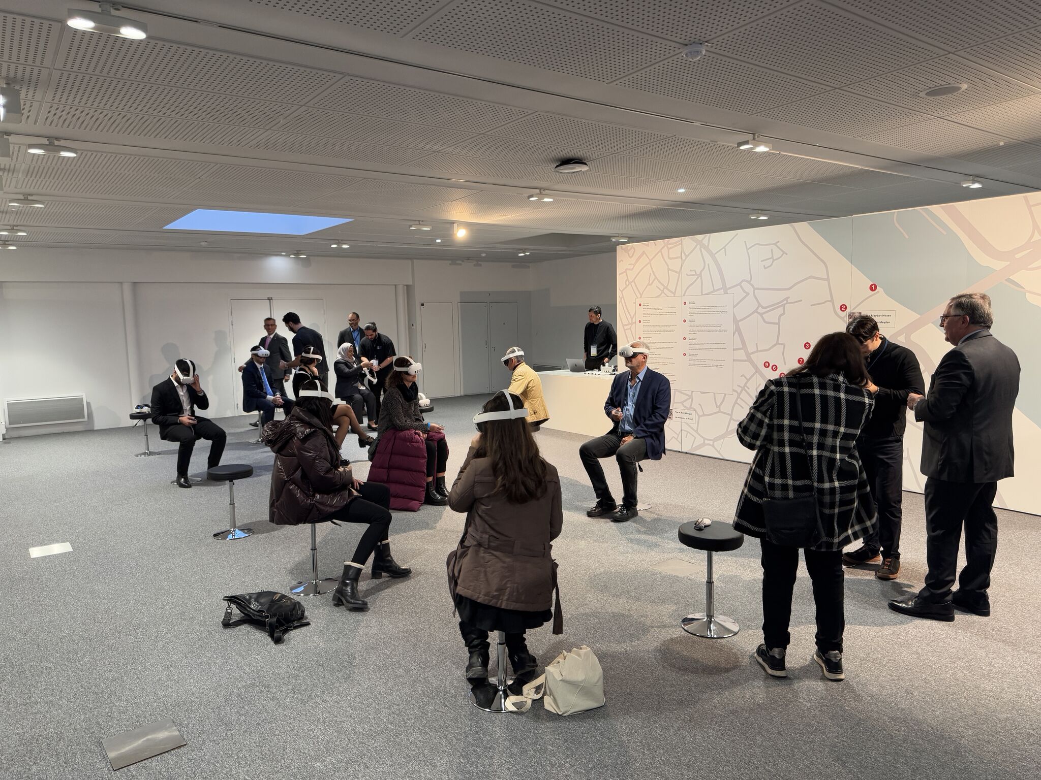 Nine people in VR headsets sit on stools at the exhibition launch, while eight people stand nearby talking.