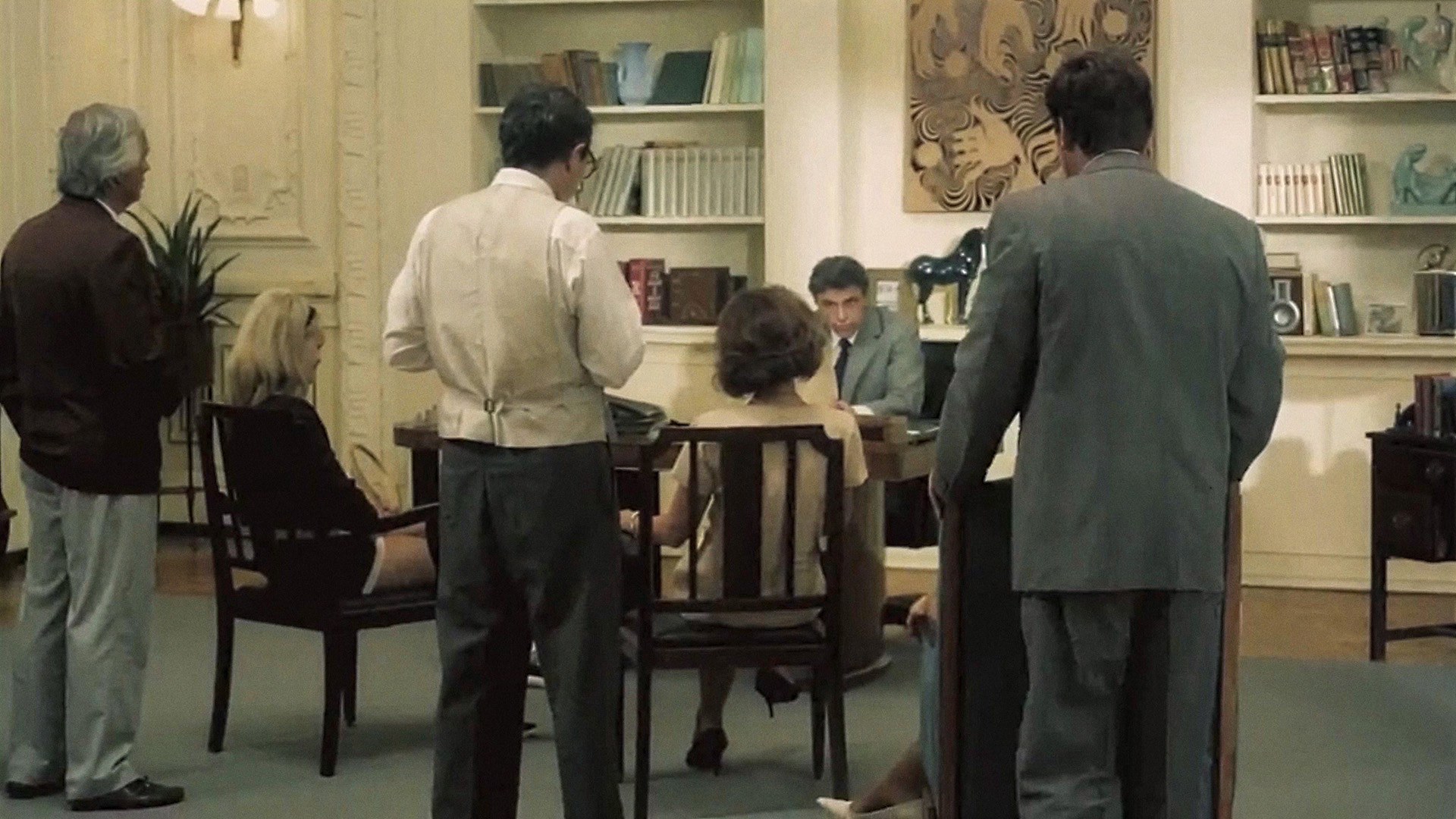 The King of Cannes: a family gathered in an ornate study, several relatives standing and seated around a desk where a man reads from documents, bookshelves lining the walls.