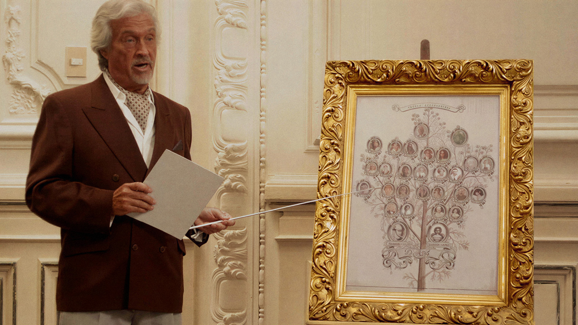 The King of Cannes: an older man in a brown suit points a stick at an ornate gold framed family tree portrait, presenting the lineage to the room.