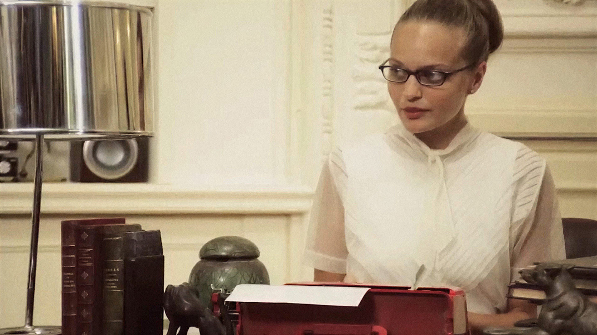 The King of Cannes: a woman in glasses sits at a desk with a vintage red typewriter, surrounded by leather bound books and a bronze dog figurine.