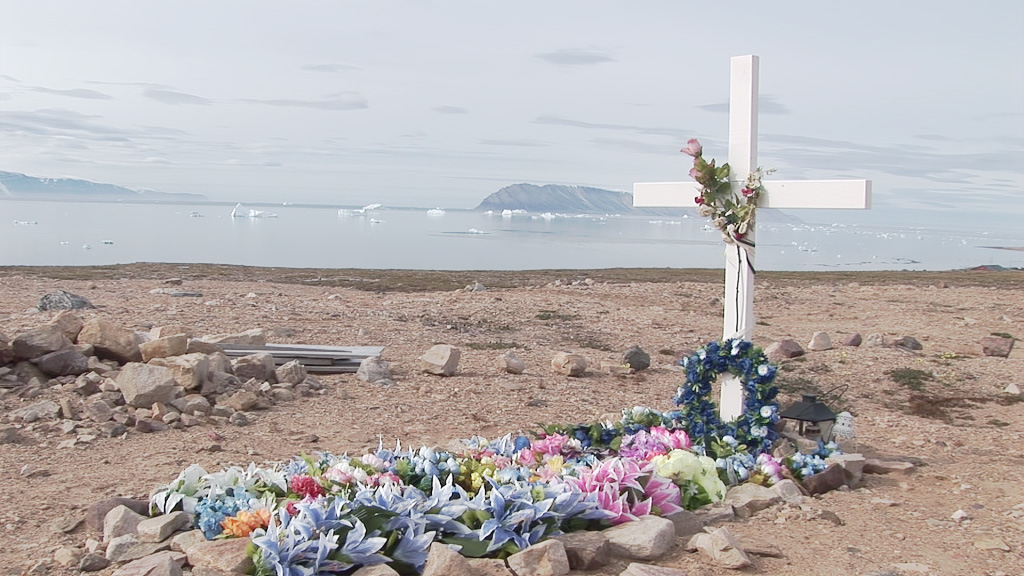 A screenshot of a cross sitting above a grave with flowers laid out around it