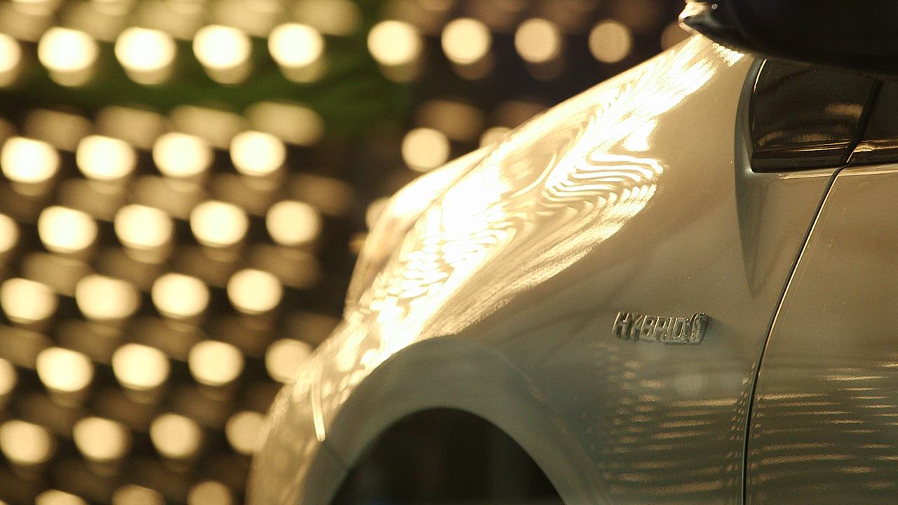 Close-up of a silver Toyota Prius fender with a Hybrid badge, reflecting a blurred grid of warm lights in the background.