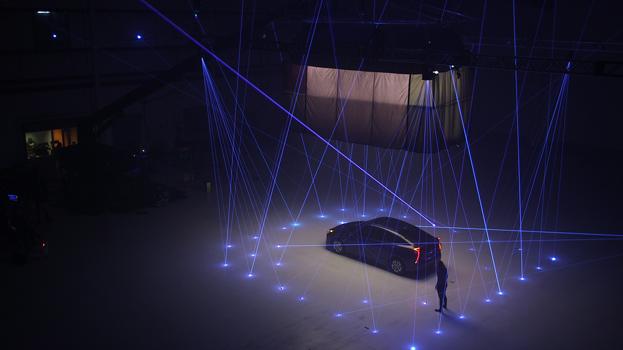 A dark car parked in an aircraft hangar surrounded by a web of glowing blue laser tripwires and floor lights.