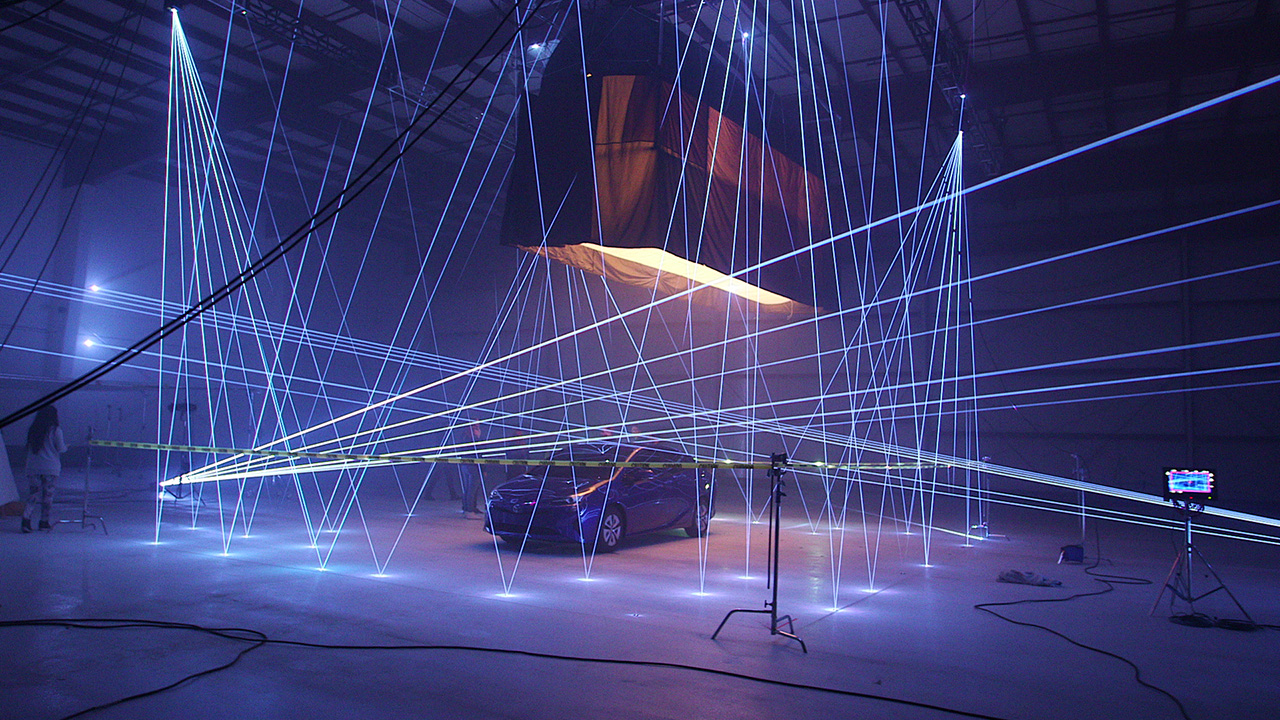 A car in a hangar beneath crossing blue laser beams and an overhead orange fabric rig, with crew and camera equipment visible nearby.
