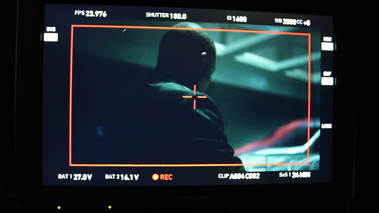A camera monitor on set displaying a framed shot of a crew member's silhouette, with recording data overlaid on screen.