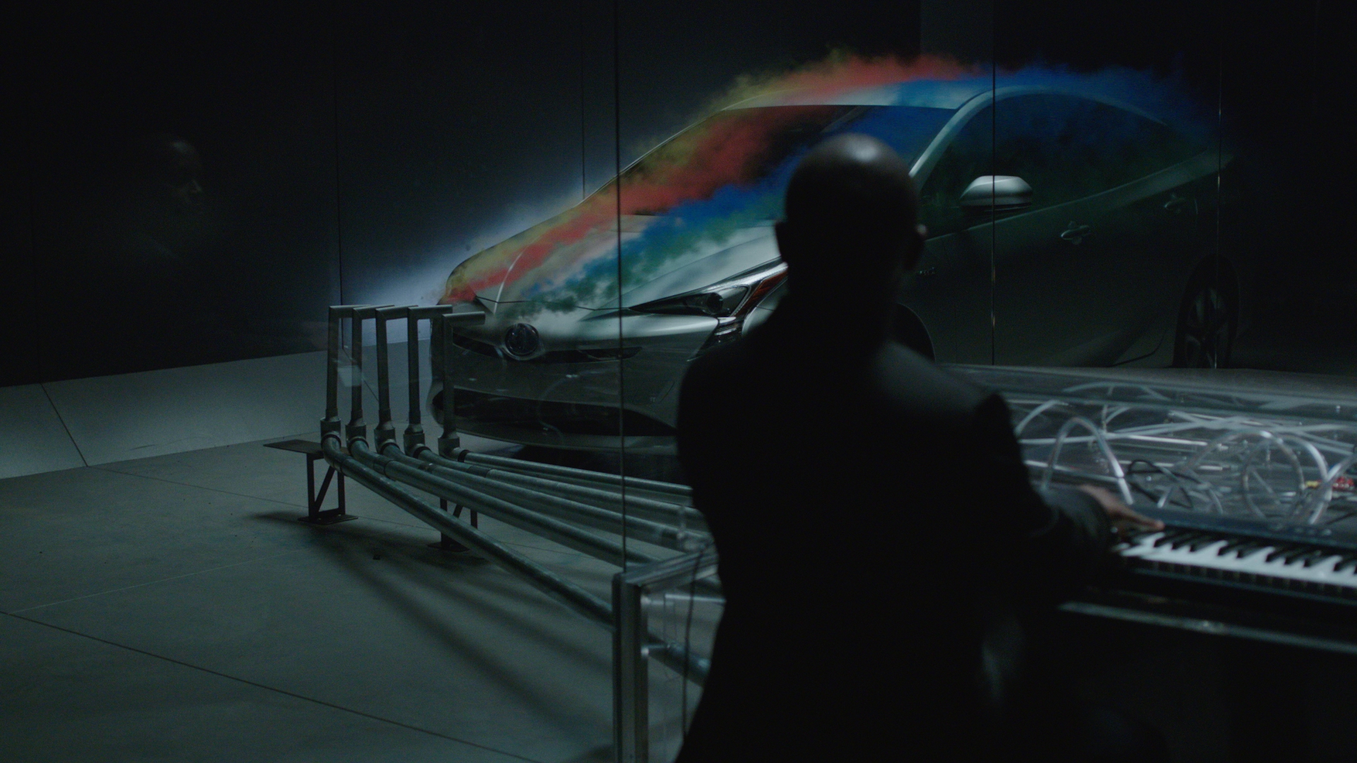A silhouetted figure plays the custom smoke-emitting acrylic piano in a darkened studio, while a vivid thermal heat-map is projected across the bonnet of the Toyota Prius inside the wind-tunnel chamber behind him.