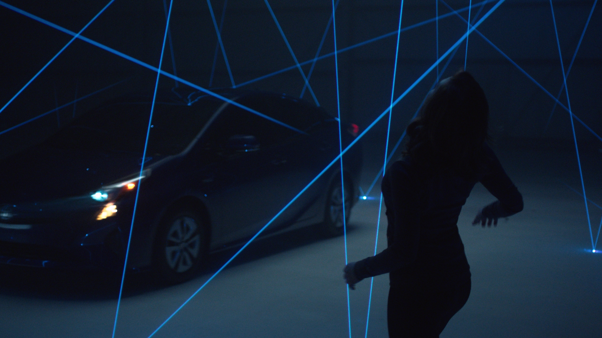 A dancer silhouetted in dark clothing moves through a web of blue laser tripwires in a darkened studio, with the Toyota Prius lit by its headlights in the background — the sensor awareness experiment.