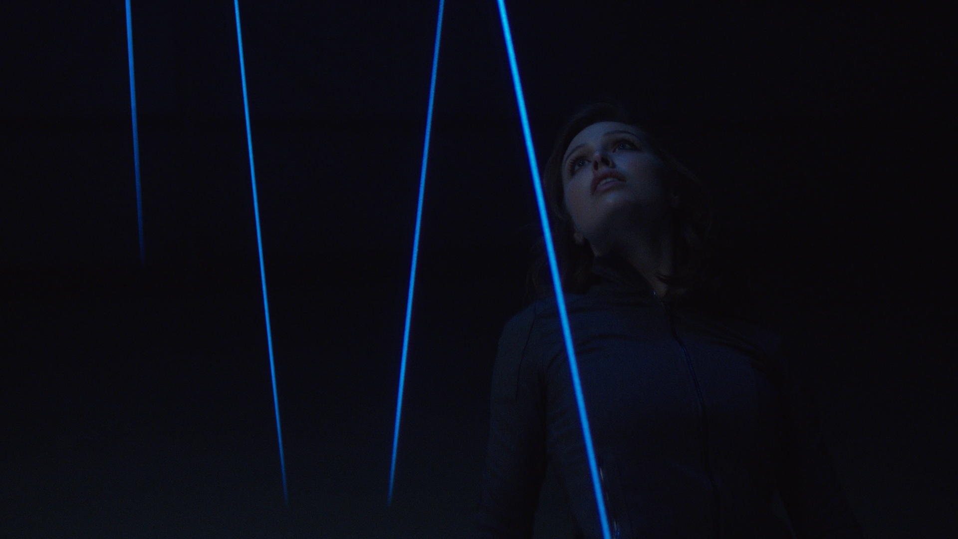 Close portrait of a woman gazing upward in near-total darkness, her face illuminated only by bright blue laser beams cutting diagonally around her during the Prius laser tripwire sequence.