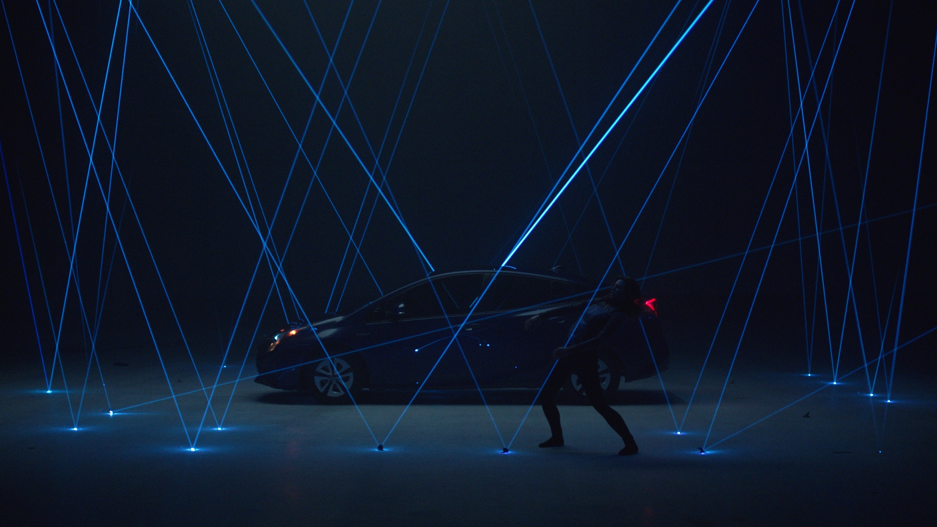 Wide cinematic shot of the Toyota Prius surrounded by a dramatic fan of blue laser beams rising from floor-mounted light points, with a dancer crouching in silhouette in front of the car.