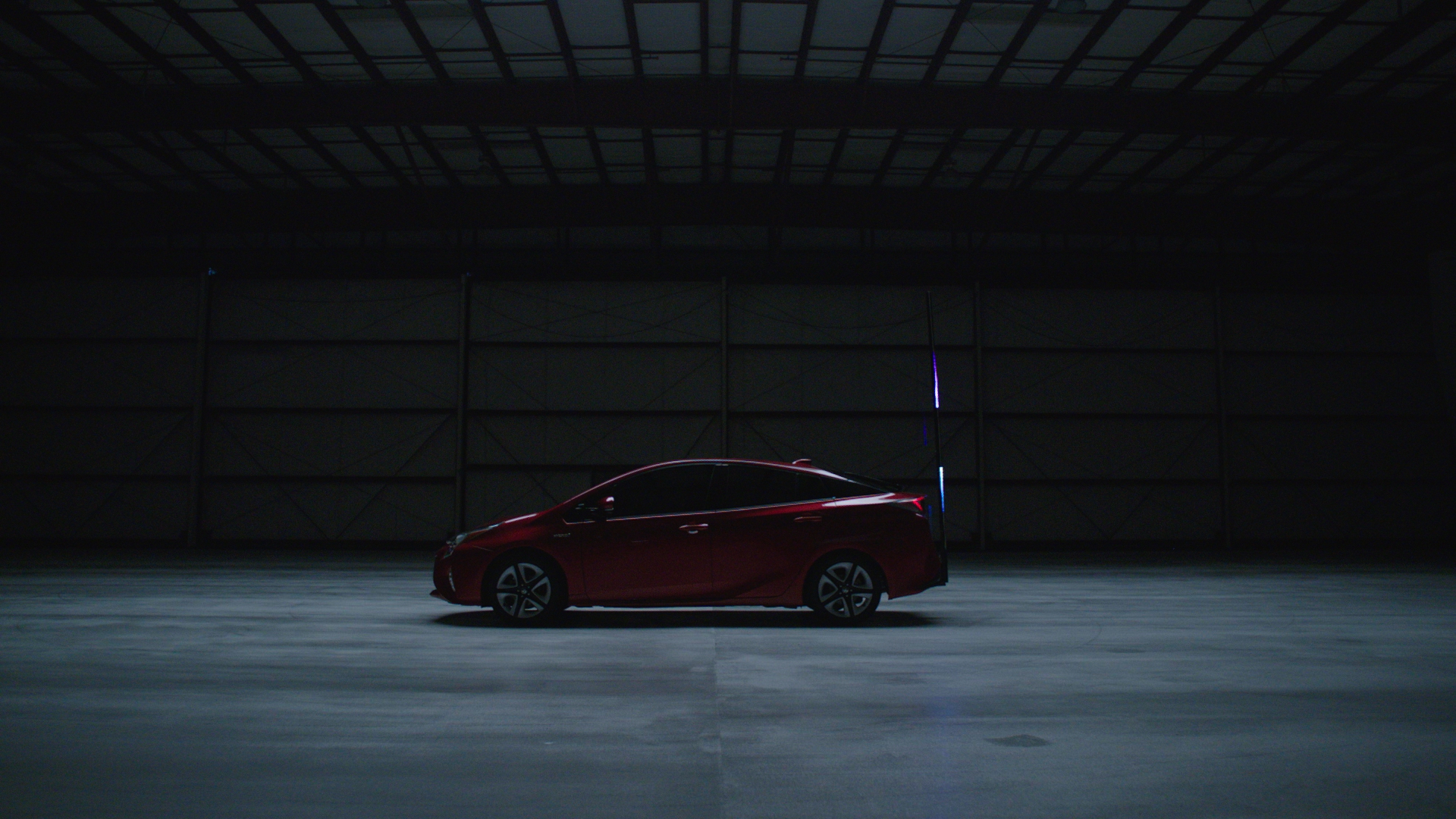Side profile of the red Toyota Prius parked alone in a dimly lit aircraft hangar, a single vertical blue laser beam glowing beside the car.