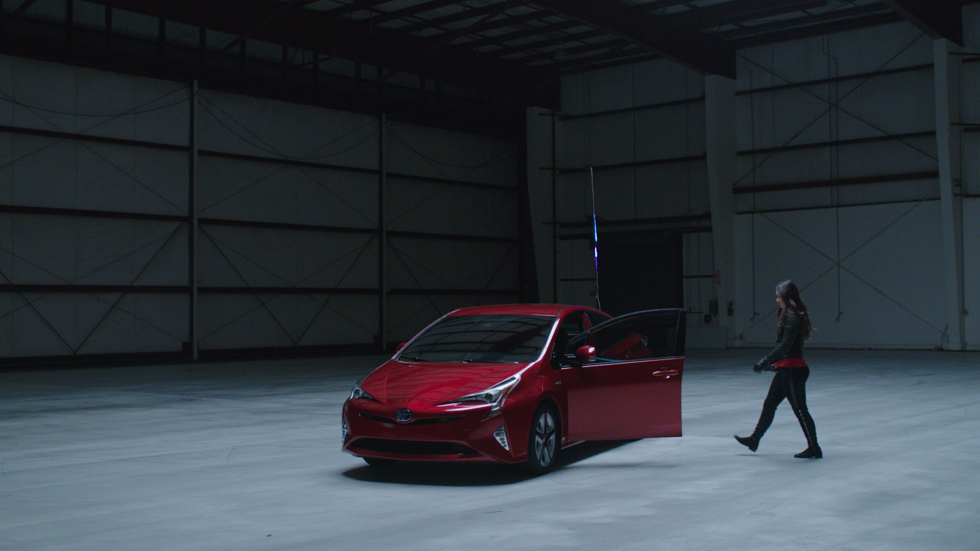 The red Toyota Prius with its driver's door open in a large aircraft hangar, as a performer dressed in dark clothing walks toward it.