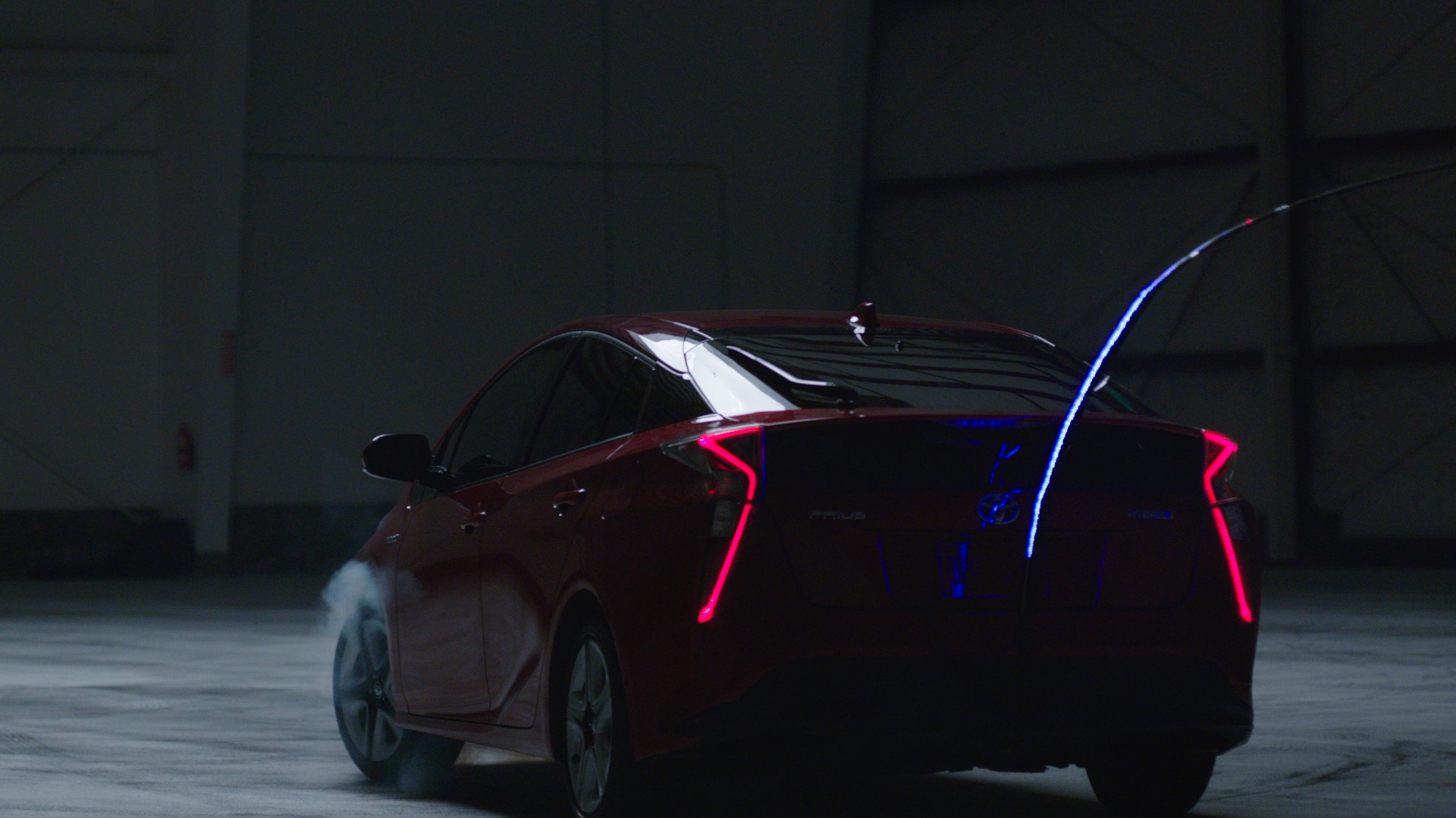 Rear three-quarter view of the red Toyota Prius with glowing pink tail lights and a blue laser arc rising from its roofline, light smoke drifting near the rear wheels in a darkened aircraft hangar.