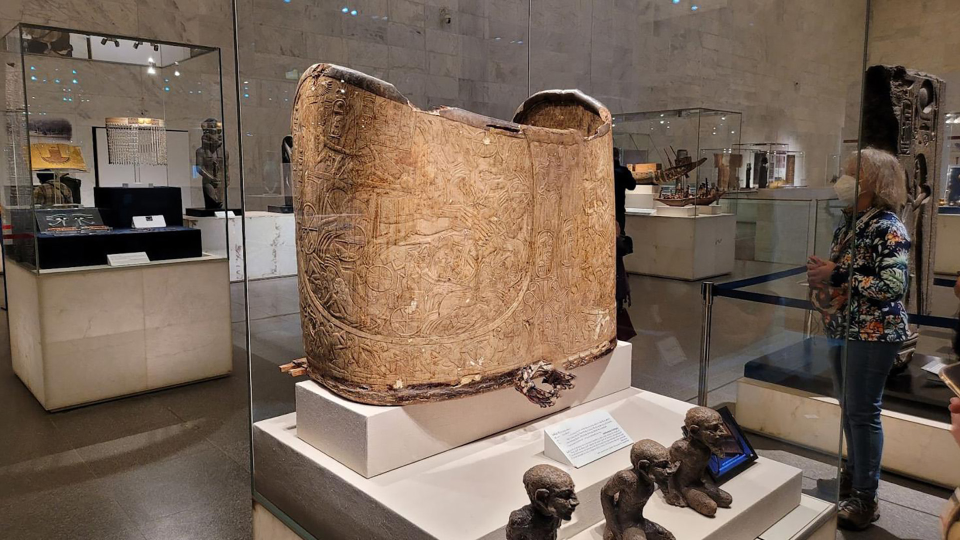 A large, hieroglyph-carved curved wooden chariot body panel displayed upright in a museum gallery, with small carved monkey figurines in the foreground and a visitor observing nearby exhibits.