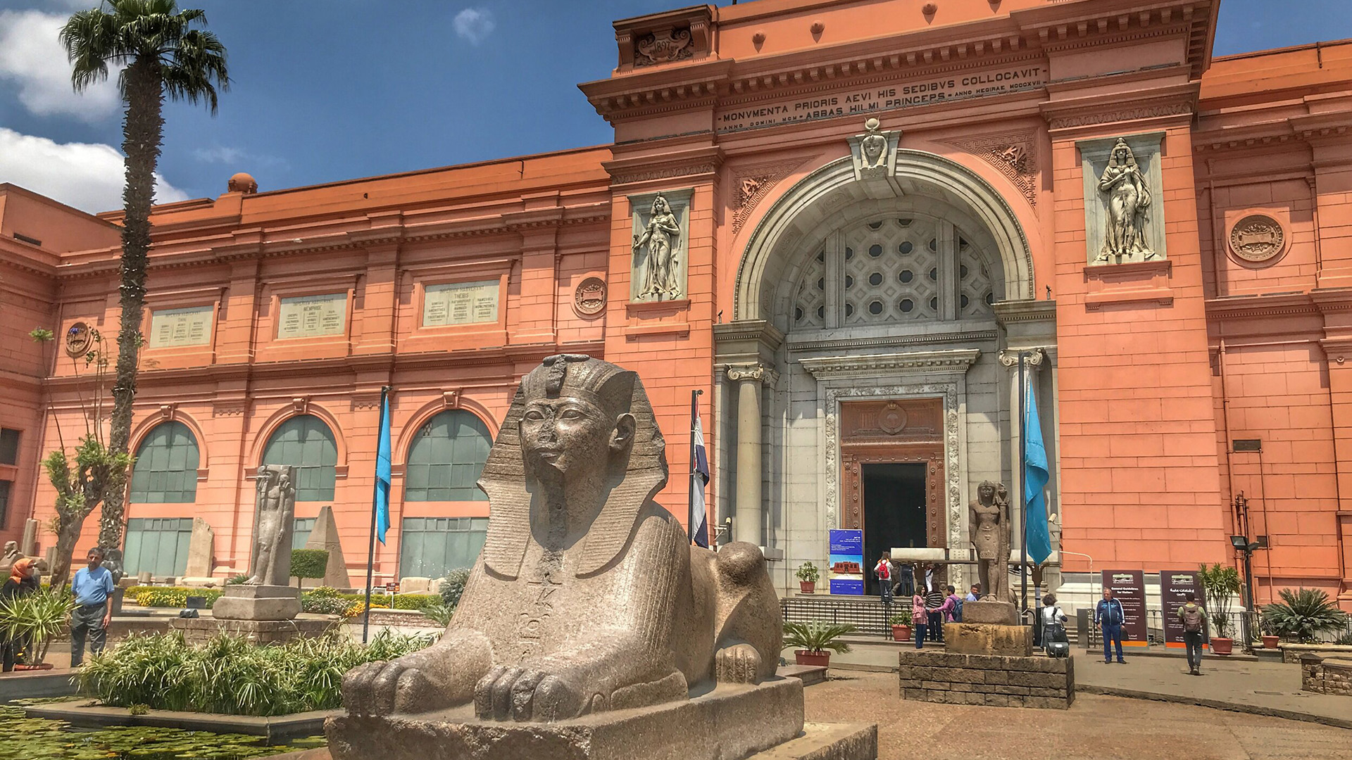The entrance of the Egyptian Museum in Cairo, with a granite sphinx statue in the foreground and visitors near the main doorway.