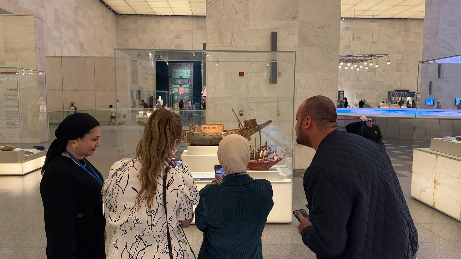 A group of visitors gather around a display case to view a model wooden boat, with a phone raised to scan it.