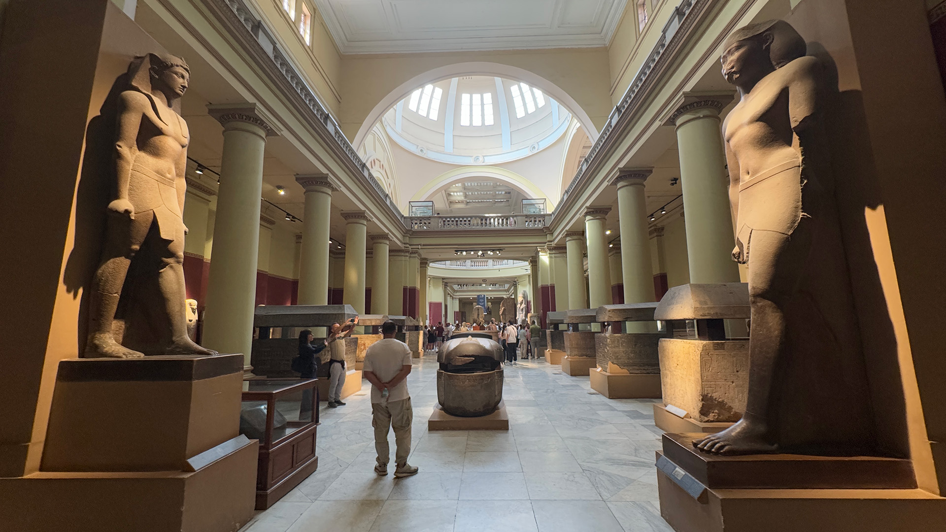 A grand museum hall lined with colossal ancient Egyptian statues and stone sarcophagi, with visitors walking through.
