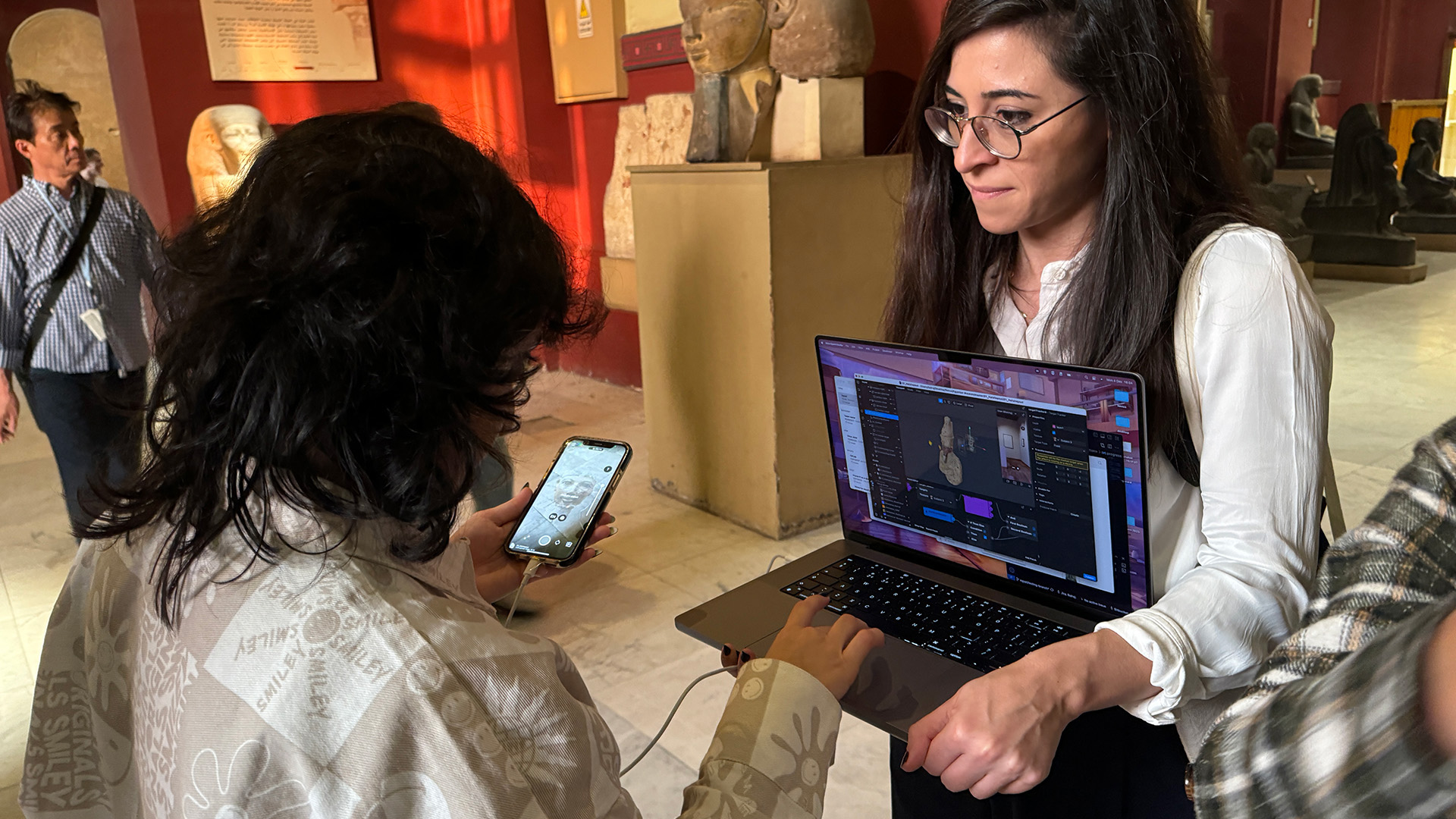 Two developers review a 3D model of a museum artefact on a laptop, with a phone held up beside it in the gallery.