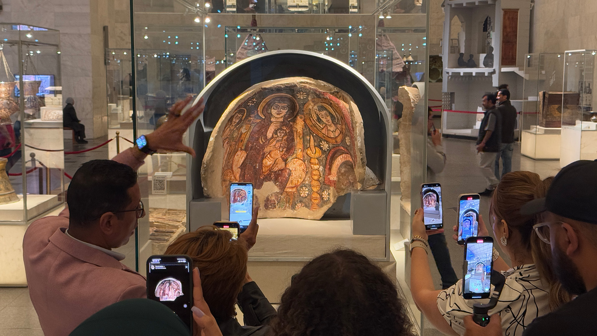 Museum visitors hold up phones running the AR app in front of a painted Coptic wall fragment displayed in a glass case.