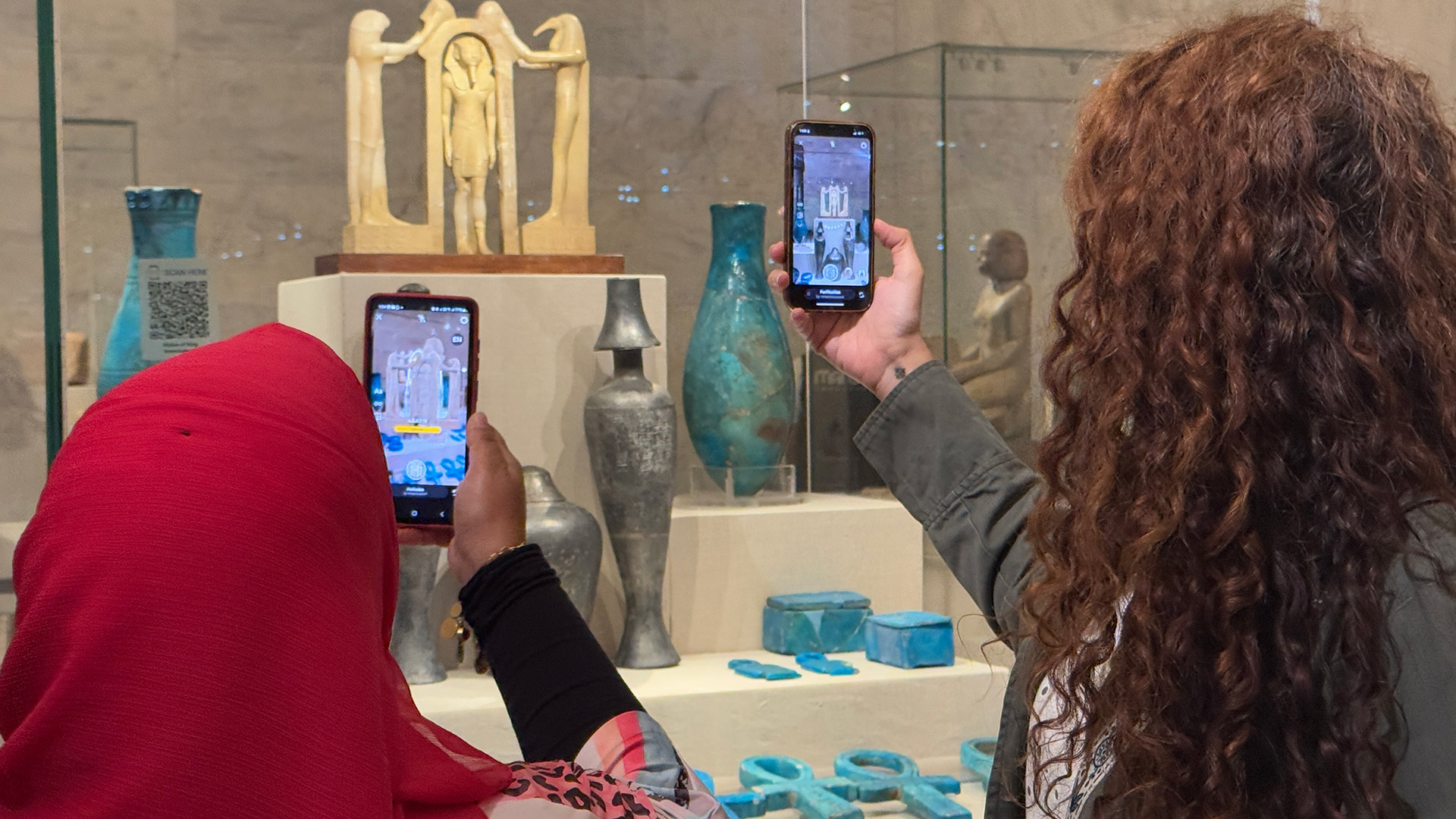 Two visitors use the AR app on their phones to view a small gilded triad statue displayed among ancient glass vessels.