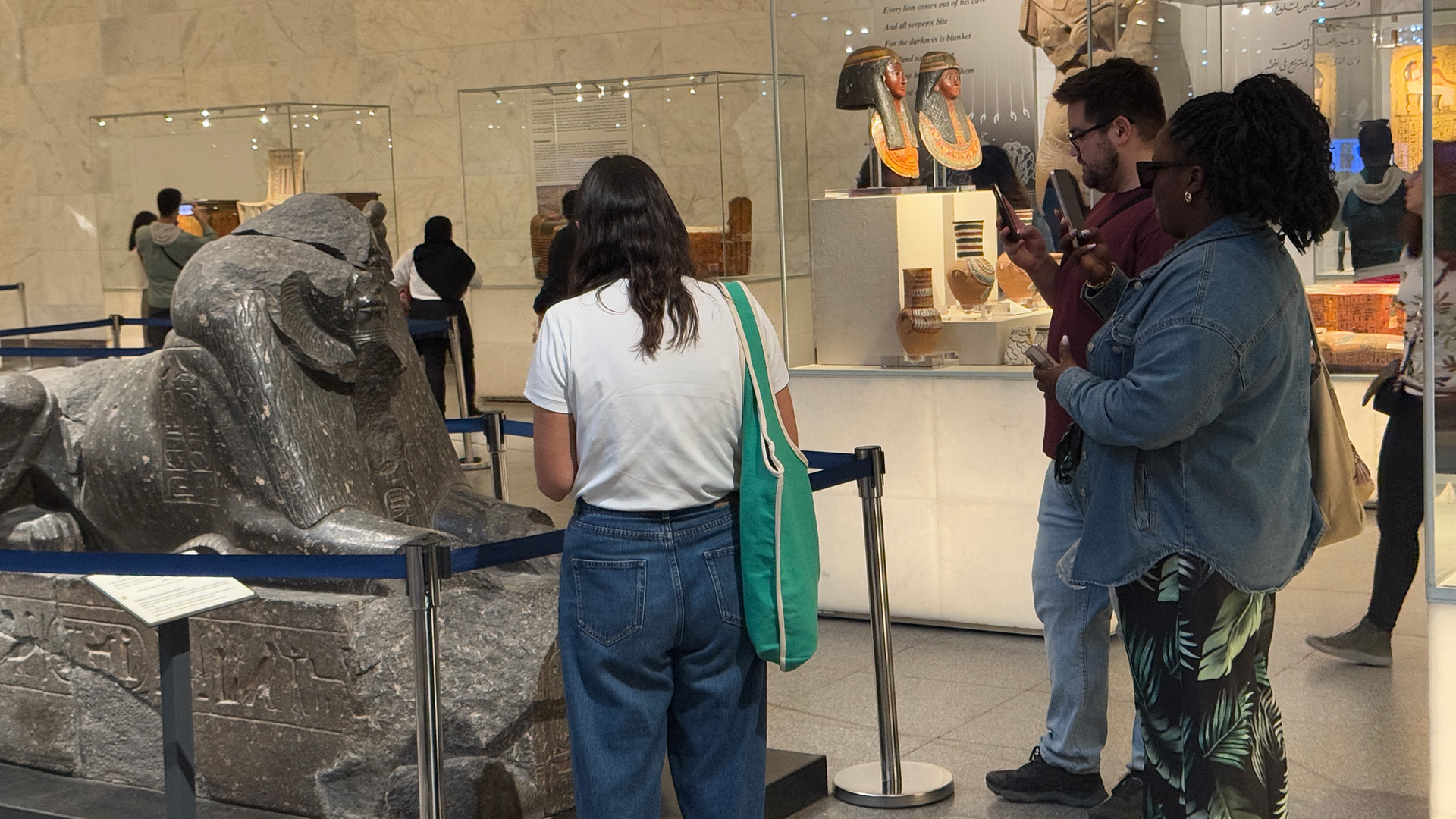 Visitors use their phones near a ram-headed sphinx statue while others photograph painted mummy cases in a nearby case.