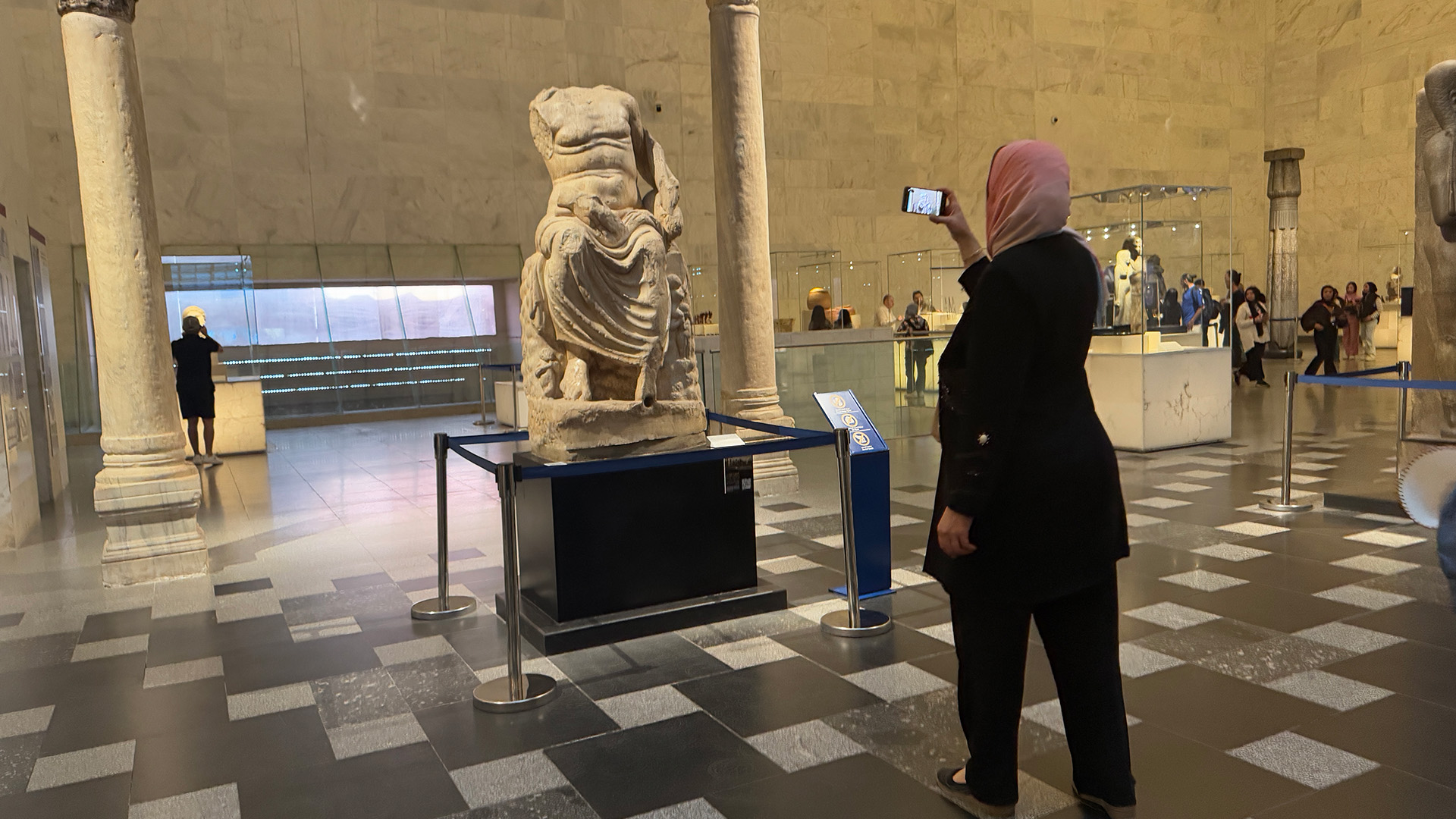A visitor in a headscarf uses the AR app on her phone to view a headless classical statue in a museum hall.