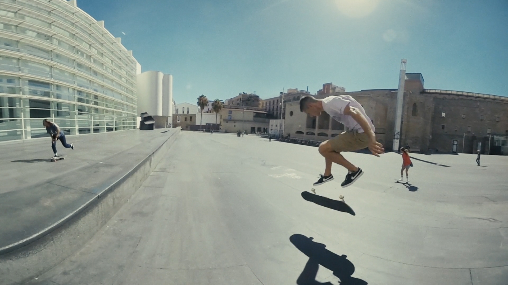Wide urban plaza with skateboarders in motion under a clear blue sky. In the foreground, a skater wearing a white T-shirt, khaki shorts, and black sneakers performs a kickflip, his shadow stretching across the pavement. Behind him, other skaters ride along the smooth concrete, with modern glass buildings on one side and historic stone architecture on the other, capturing the blend of city culture and skateboarding energy.