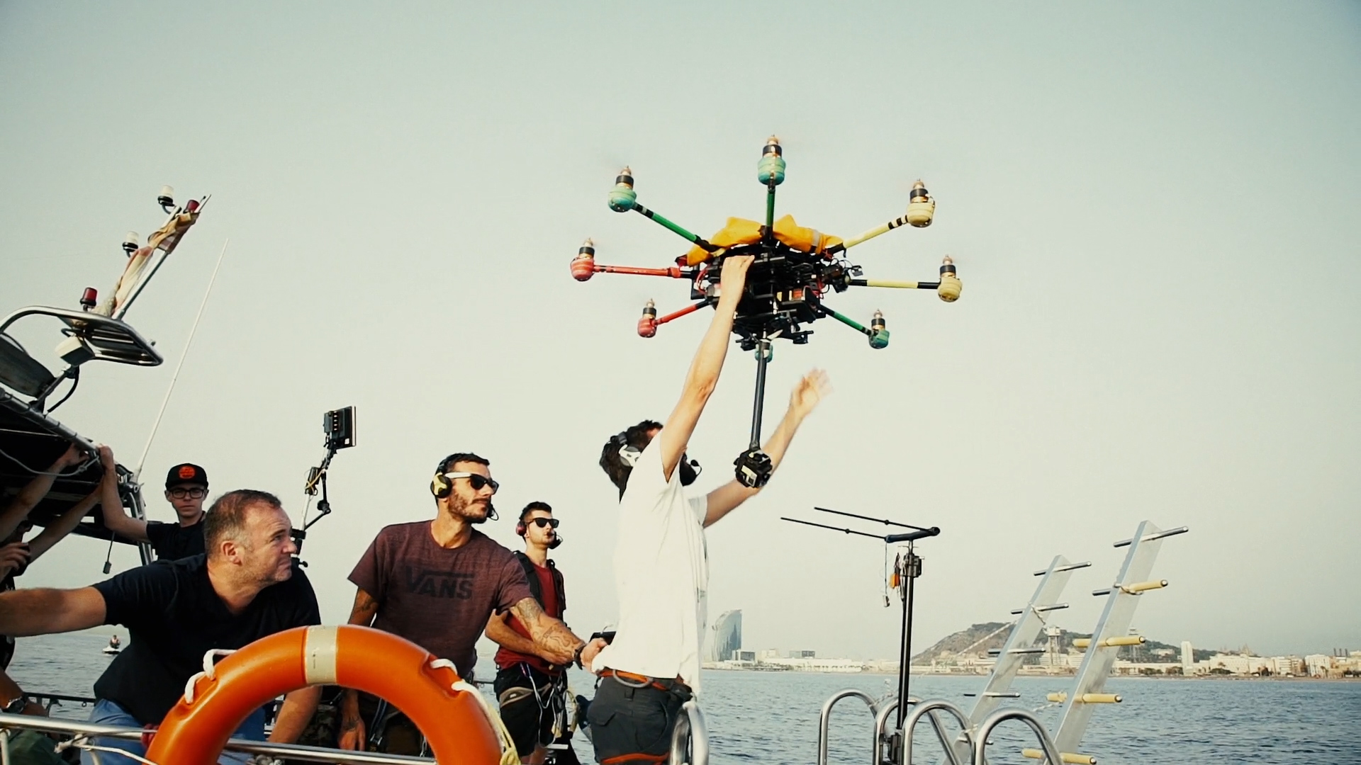 Film crew on a boat preparing to launch a large multi-rotor drone over the sea in Barcelona. One crew member holds the drone overhead while others stand by with headsets, cameras, and safety gear. The city’s skyline and W Hotel tower are visible in the background, blending filmmaking technology with the Mediterranean coastal setting.