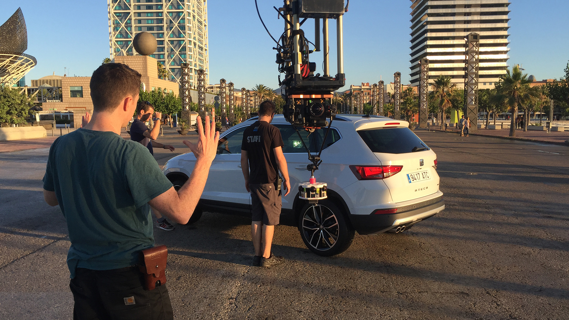 Film crew in Barcelona preparing a car-mounted camera rig on a white SEAT Ateca SUV during a shoot. A large stabilized camera system is fixed to the vehicle’s side while crew members guide the setup, with one person directing by raising their hand. The background shows Barcelona landmarks including the twin towers near Port Olímpic and Frank Gehry’s golden fish sculpture, captured in the warm light of late afternoon.