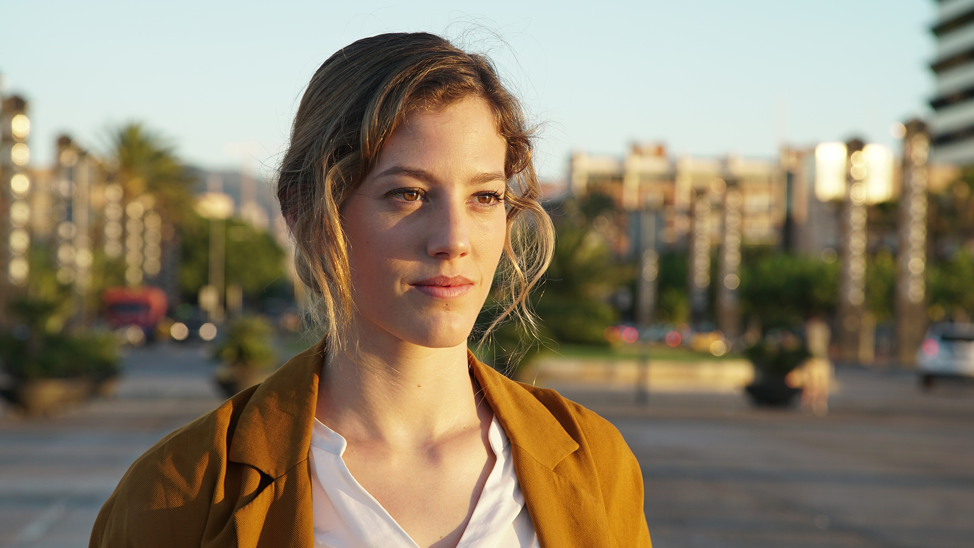 Portrait of stuntwoman Natasha Duran standing outdoors in Barcelona during golden hour. She wears a mustard-colored jacket over a white shirt, her hair loosely tied back, and she gazes forward with a calm, focused expression. The background is softly blurred with palm trees, city lights, and urban structures, highlighting her as the subject against the warm evening glow.