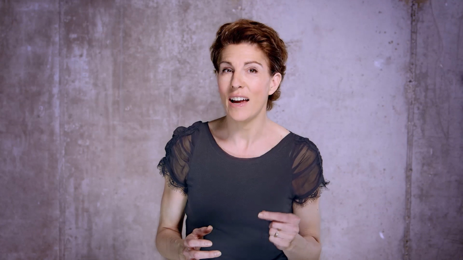 A woman with short auburn hair, wearing a black lace-sleeve top, speaking and gesturing during a filmed video segment against a bare concrete wall.