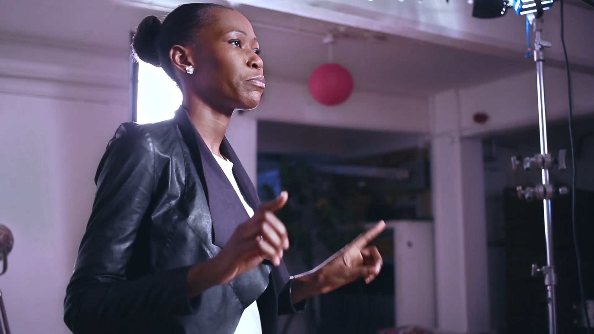 A woman with short dark hair in a bun, wearing a black leather jacket, gesturing mid-speech in a room with a pink pendant light and film lighting stands visible behind her.