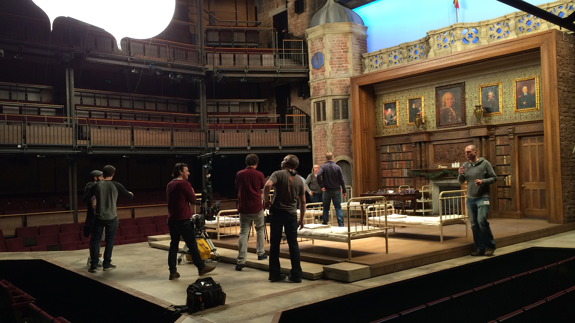Wide view of a theatre stage set featuring a brick clock tower, a library and fireplace backdrop with portrait paintings, iron-frame beds, and film crew and camera equipment in the foreground.