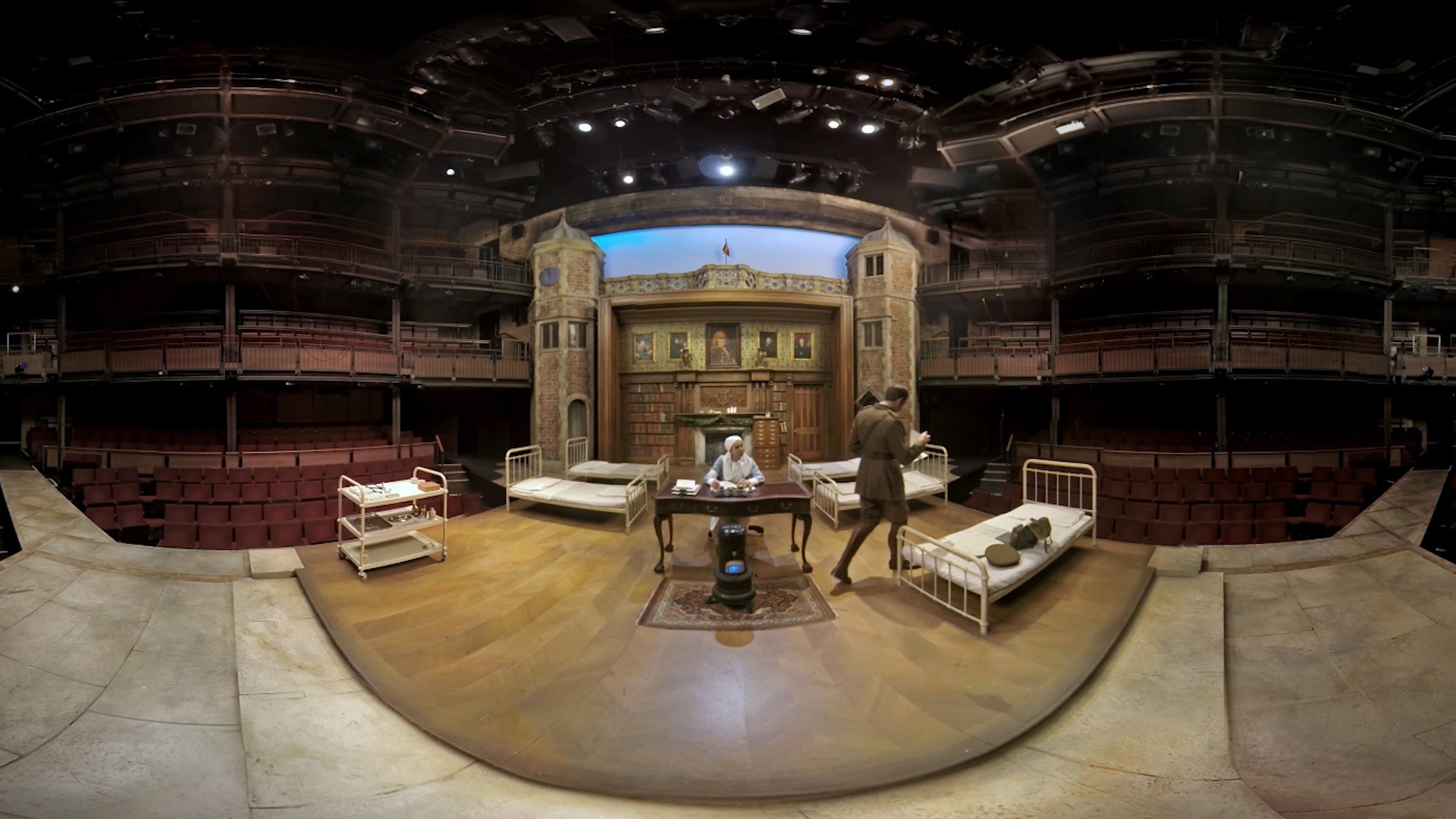 Two hands holding a Samsung tablet displaying a 360° VR view of the theatre stage set, with the brick clock tower and library backdrop visible on screen.