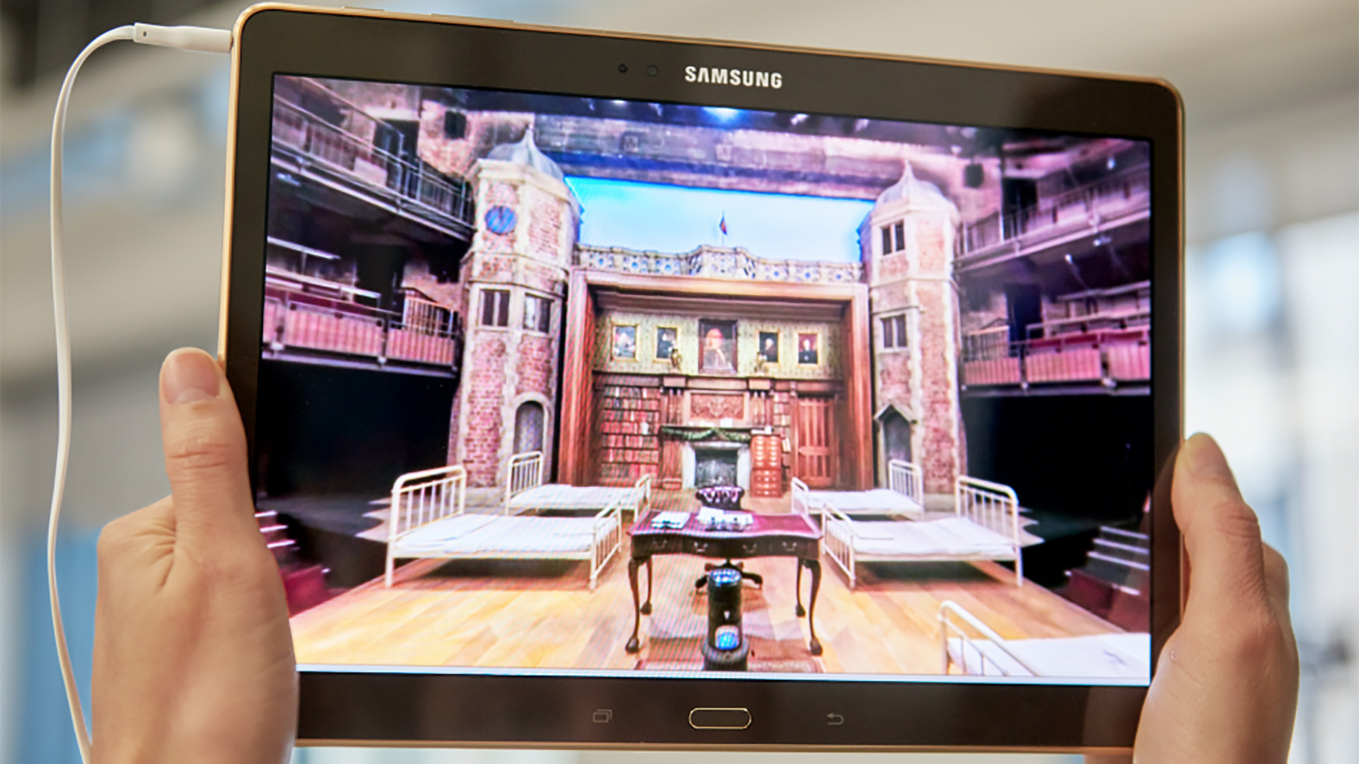 Two hands holding a Samsung tablet showing the app's 360° VR panorama of the theatre set, with the clock tower and fireplace library visible on screen.